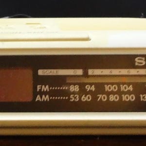 May include: A cream-colored Sony FM/AM clock radio. The display shows AM and PM indicators. The radio has a frequency dial with FM and AM settings. The model number is ICF-C25.