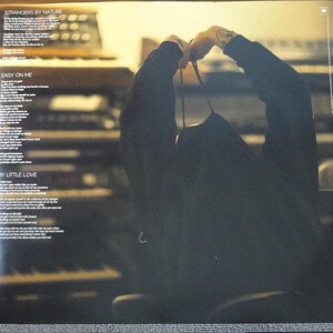May include: A dark album cover with song lyrics and the titles "Strangers by Nature", "Easy on Me", and "My Little Love". A person is holding the album. The background is a blurred image of a keyboard.