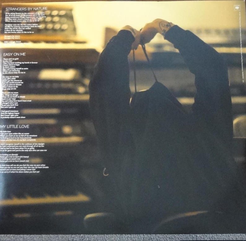 May include: A dark album cover with song lyrics and the titles "Strangers by Nature", "Easy on Me", and "My Little Love". A person is holding the album. The background is a blurred image of a keyboard.