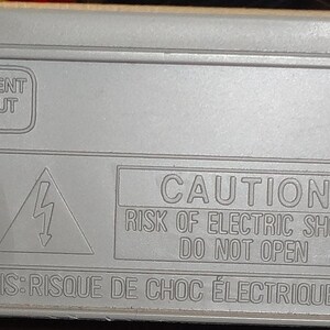 May include: The rear of a grey DVD player with various input and output ports, including RCA, S-Video, and a coaxial cable connector. The player has a warning label that reads "CAUTION RISK OF ELECTRIC SHOCK, DO NOT OPEN".