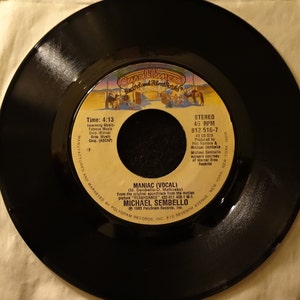 May include: Black and white 45 RPM vinyl record with the title "MANIAC (VOCAL)" by Michael Sembello. The record is from the soundtrack of the movie "FLASHDANCE".