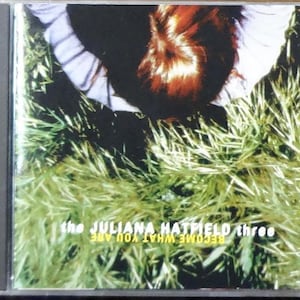 May include: CD case featuring the album "Become What You Are" by The Juliana Hatfield Three. The cover art shows a person lying in grass, with the band's name and album title in white text.
