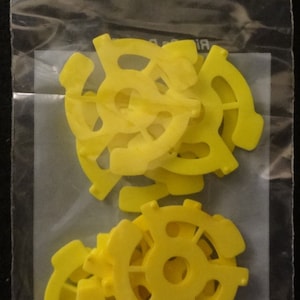 May include: A clear plastic bag containing a collection of bright yellow plastic record adapters. The adapters have a circular design with a central hole and curved arms extending outwards. The bag is sealed at the top and has a small hole for hanging.