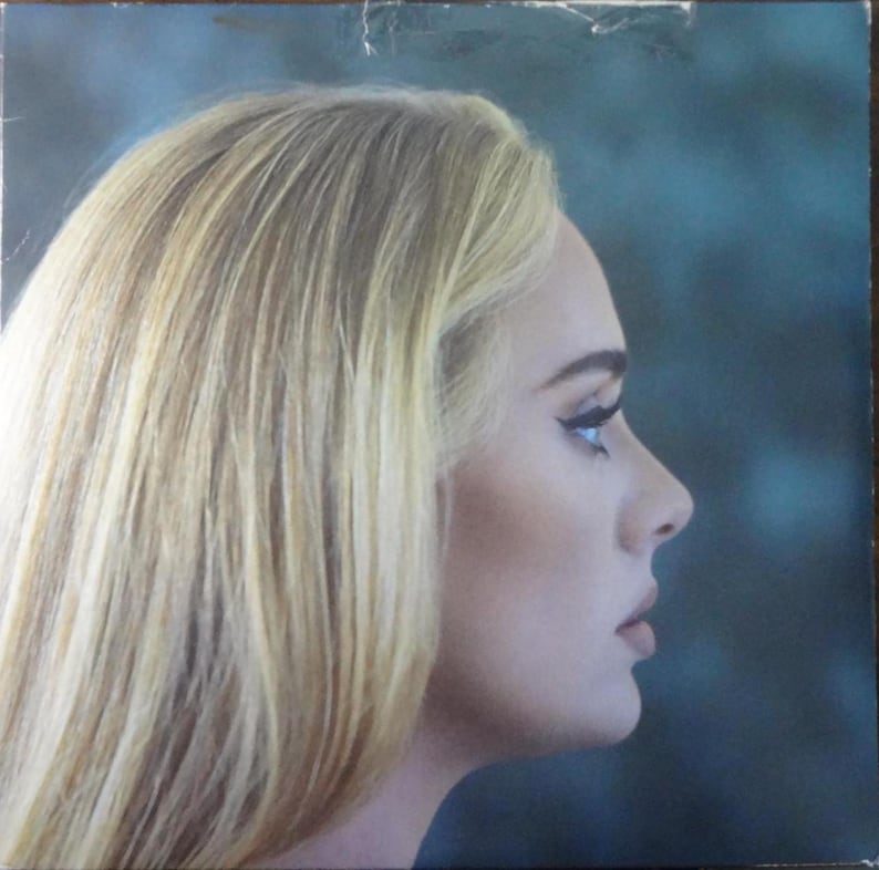 May include: Album cover featuring a side profile of a person with blonde hair and a fair complexion. The person is looking to the right, with a dark eyeliner and a neutral lip color. The background is a gradient of blues and greens.