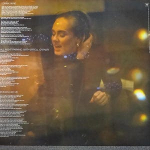 May include: Album cover featuring a woman with her hand near her head, with song lyrics visible. The album title is not visible. The background is dark with blurred lights. The album's song titles include "I Drink Wine", "All Night Parking (with Erroll Garner)", and "Woman Like Me".