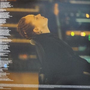 May include: A vinyl record album cover featuring a woman with auburn hair looking upwards. She wears a black top and gold hoop earrings. The background is blurred with city lights. Song lyrics are printed on the left side of the album cover.