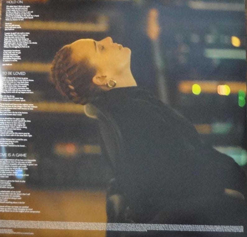 May include: A vinyl record album cover featuring a woman with auburn hair looking upwards. She wears a black top and gold hoop earrings. The background is blurred with city lights. Song lyrics are printed on the left side of the album cover.