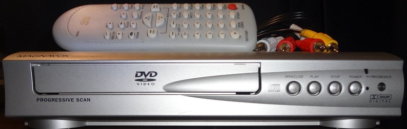 May include: A silver DVD player with a silver remote control. The DVD player has a slot for a disc, buttons for open/close, play, stop, power, and progressive scan. The remote control has buttons for various functions, including play, stop, and rewind.