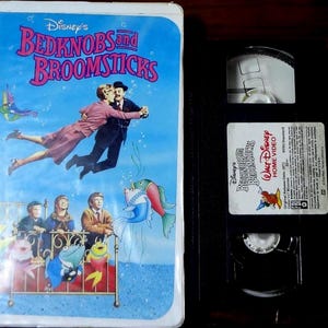 May include: A Disney Bedknobs and Broomsticks VHS tape with a white plastic case and a black cassette. The cover features a blue background with a flying couple, three children, and colorful cartoon fish. The cassette label includes movie details.