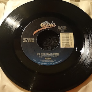 May include: A black vinyl record with a white label. The label has the text "Epic" in a stylized font, the song title "99 Red Balloons", the artist "Nena", and the songwriters and producers. The record is a 45 RPM record.
