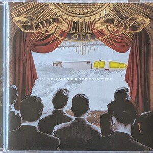 May include: A Fall Out Boy CD titled From Under the Cork Tree. The cover art depicts a group of people in dark suits viewed from behind, looking toward a stage with red curtains. A snowy landscape with a white trailer and yellow van is visible.