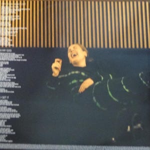 May include: Image of a person laughing, wearing a black top with green accents. The background features vertical wooden slats and song lyrics. The image is dark with a focus on the person's expression.