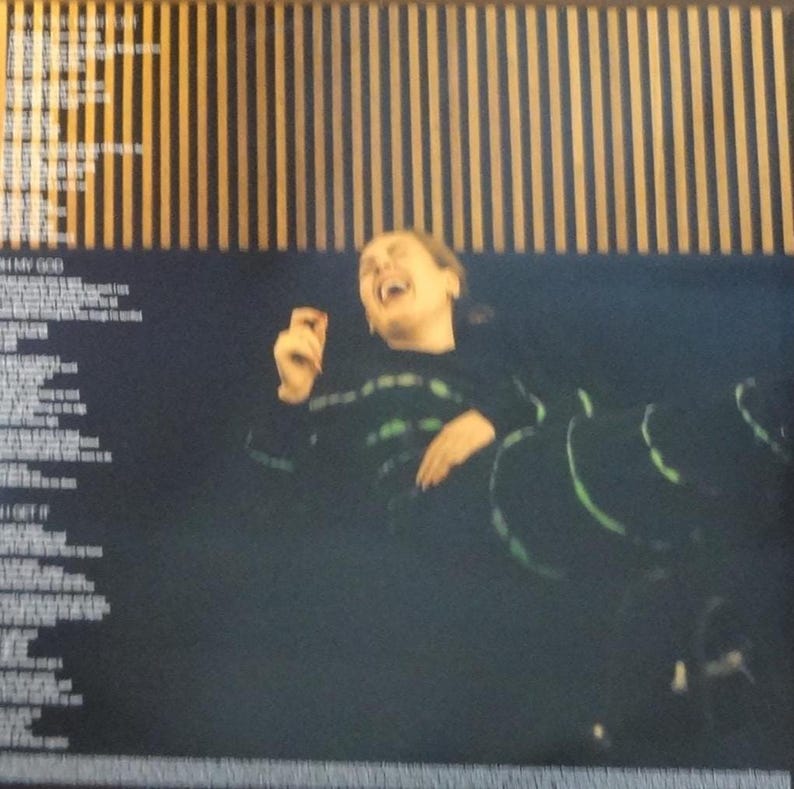 May include: Image of a person laughing, wearing a black top with green accents. The background features vertical wooden slats and song lyrics. The image is dark with a focus on the person's expression.