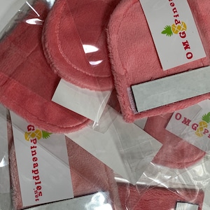 Op de afbeelding: Roze pluche vierkante stoffen met een wit label waarop "OMG Pineapples" staat en een ananasafbeelding. De stoffen vierkanten hebben een witte kleefrug.