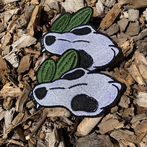 May include: Two embroidered patches featuring a white skull with black eyes and a green leaf design. The patches are on a brown wood chip background.