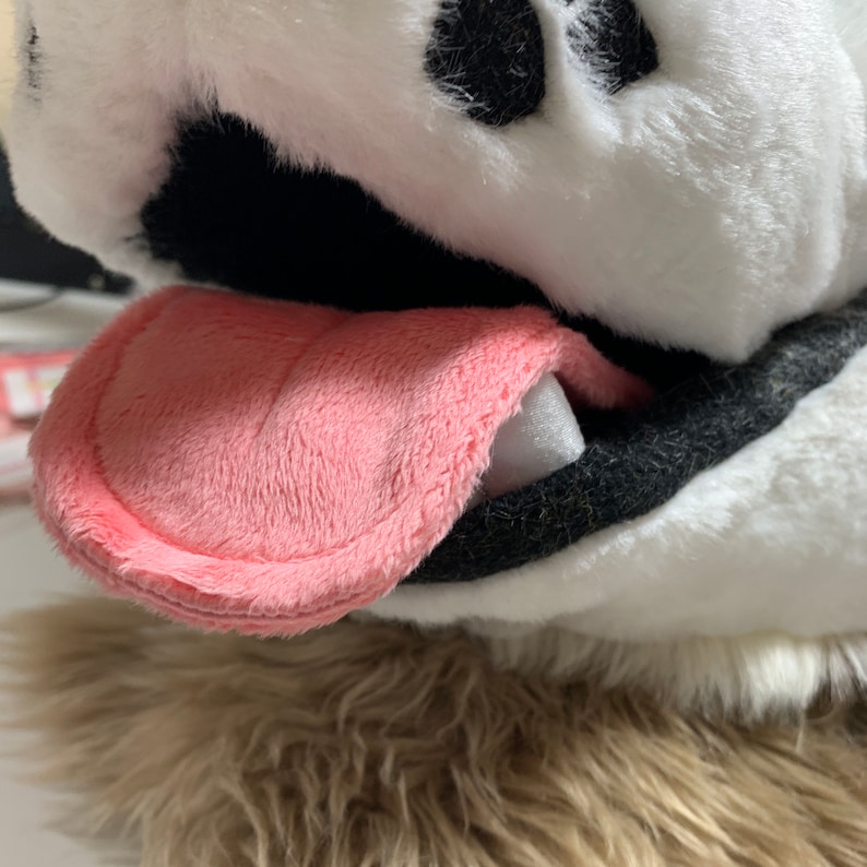 Op de afbeelding: Close-up van de bek van een pluche hond met een roze tong die eruit steekt. De vacht van de hond is wit en zwart, en de tong is gemaakt van zacht, pluche stof.