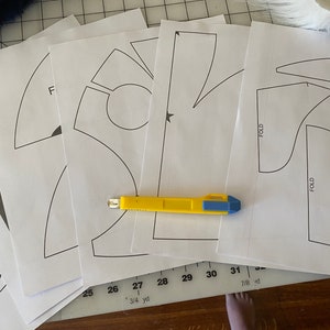 May include: A set of five white paper patterns for sewing, with a yellow utility knife on top. The patterns include a variety of shapes, including a star, a circle, and a curved line. The patterns are labeled with numbers and measurements in inches.