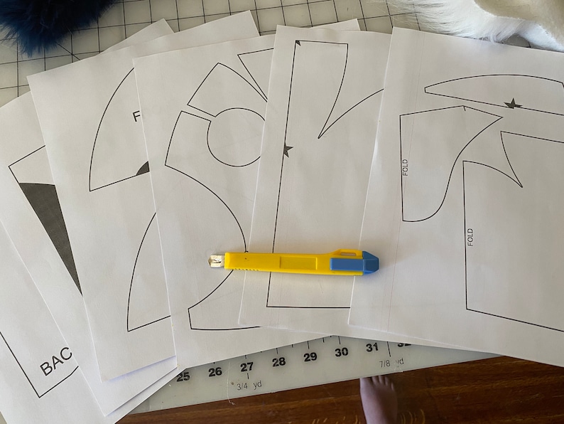 May include: A set of five white paper patterns for sewing, with a yellow utility knife on top. The patterns include a variety of shapes, including a star, a circle, and a curved line. The patterns are labeled with numbers and measurements in inches.