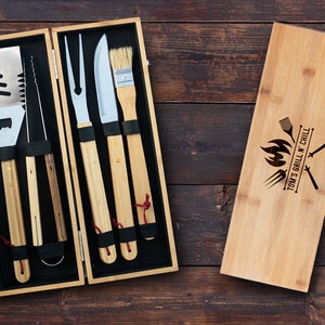 May include: A bamboo grilling tool set with a wooden carrying case. The tools include a spatula, tongs, a fork, a knife, and a basting brush. The case is engraved with the text "Tom's Grill N Chill" and a grill-themed design.