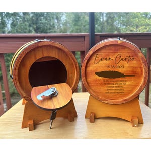 May include: Two wooden barrel-shaped boxes with lids. The box on the left is open and has a keyhole and a metal latch. The box on the right is closed and has a personalized inscription that reads "Evan Carter 1978-2023" with a feather design and a quote about remembering a loved one.
