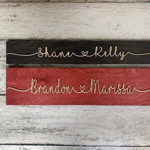 Puede incluir: Dos letreros de madera con nombres y un s&iacute;mbolo de coraz&oacute;n. El letrero superior es negro con los nombres "Shane & Kelly" en blanco. El letrero inferior es rojo con los nombres "Brandon & Marissa" en blanco.