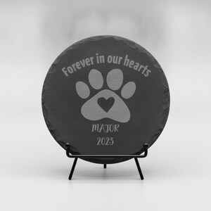 May include: A circular dark gray slate plaque with rough, uneven edges, displayed on a black metal stand. The surface features an engraved paw print with a heart in the center, the text "Forever in our hearts", the name "MAJOR", and the year "2023".