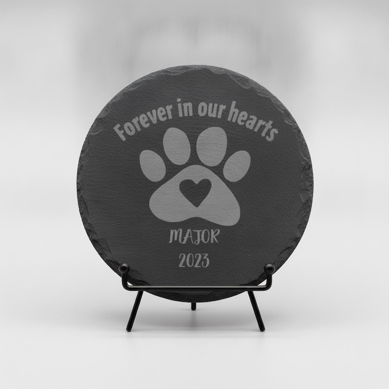 May include: A circular dark gray slate plaque with rough, uneven edges, displayed on a black metal stand. The surface features an engraved paw print with a heart in the center, the text "Forever in our hearts", the name "MAJOR", and the year "2023".