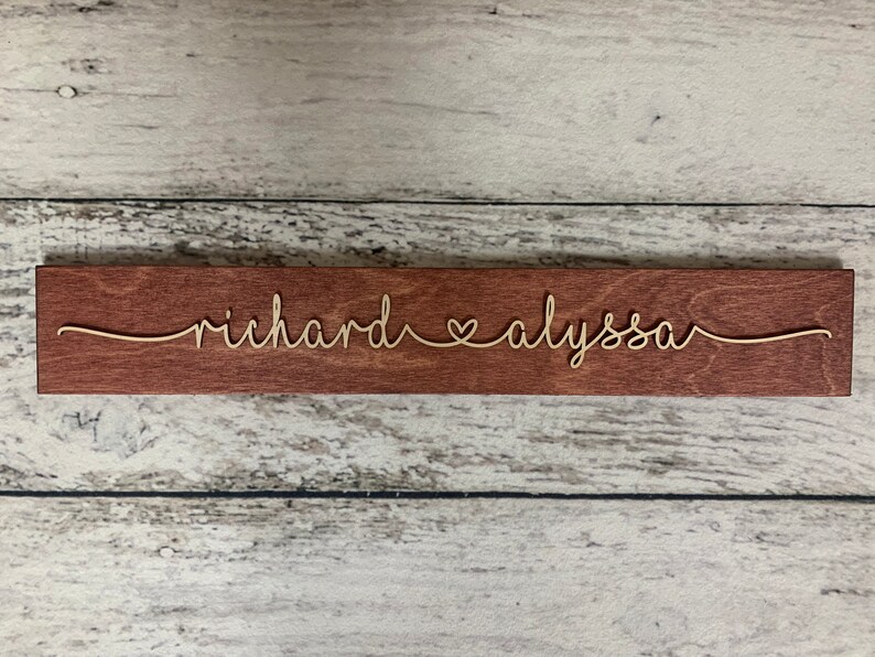 Puede incluir: Un letrero de madera con los nombres "richard" y "alyssa" escritos en cursiva con un s&iacute;mbolo de coraz&oacute;n entre ellos.