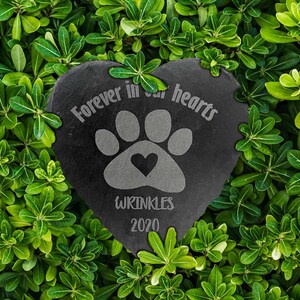 May include: A heart-shaped black slate plaque featuring an engraved paw print with a small heart in the center. The text "Forever in our hearts" is arched above the paw, with the name "WRINKLES" and the year "2020" engraved below the design.