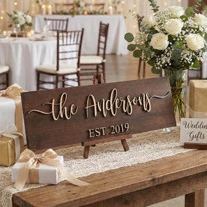 May include: A rectangular dark brown wooden sign displayed on a small wooden easel, featuring the raised light-colored text "the Andersons" in a cursive script and "EST. 2019" in a block font, positioned on a table with gift boxes and flowers.
