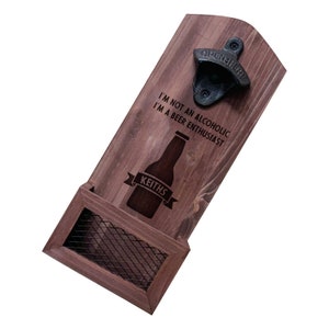 May include: Wooden wall mounted bottle opener with a metal bottle opener and a small shelf for holding bottles. The bottle opener has the text "Open Here" on it. The shelf has a wire mesh insert. The wood is a dark brown color. The text "I'm not an alcoholic, I'm a beer enthusiast" is engraved on the wood. The text "Keith's" is engraved on a banner below a bottle graphic.