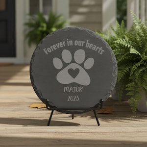 May include: A circular dark gray slate memorial plaque featuring the engraved text "Forever in our hearts" above a paw print graphic with a heart in the center, followed by the name "MAJOR" and the year "2023" on a black metal display stand.