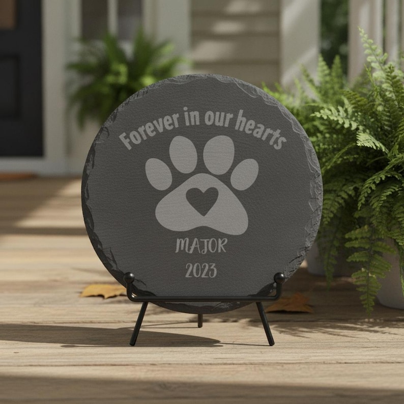 May include: A circular dark gray slate memorial plaque featuring the engraved text "Forever in our hearts" above a paw print graphic with a heart in the center, followed by the name "MAJOR" and the year "2023" on a black metal display stand.