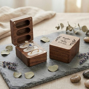 May include: A wooden ring box with three slots, each holding a ring. The box is open, revealing a gold band, a diamond ring, and a ring with a blue stone. A second box is closed, with the names "Braden & Sheila" and the date "10.14.2026" engraved on the top.