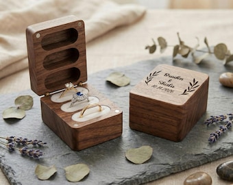 Puede incluir: Una caja de anillos de madera con tres ranuras, cada una con un anillo. La caja está abierta, revelando una alianza de oro, un anillo de diamantes y un anillo con una piedra azul. Una segunda caja está cerrada, con los nombres "Braden & Sheila" y la fecha "10.14.2026" grabados.