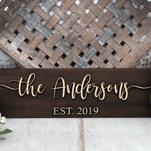 May include: A dark brown wooden sign with the text "the Andersons EST. 2019" in gold lettering.