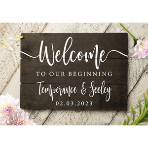 May include: A rustic wooden sign with the text "Welcome to our beginning Temperance & Seeley 02.03.2023" in white lettering.