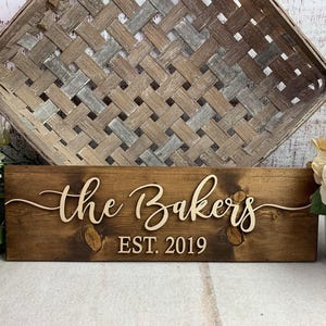 May include: A rectangular dark brown wooden sign featuring the raised white script text the Bakers and the block text EST. 2019. The sign is positioned in front of a large woven basket and flanked by white and cream artificial flowers.