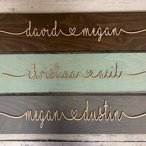 Puede incluir: Tres letreros de madera con nombres y un s&iacute;mbolo de coraz&oacute;n en escritura cursiva. El letrero superior es marr&oacute;n oscuro con los nombres "david" y "megan". El letrero del medio es azul claro con los nombres "christian" y "neil". El letrero inferior es gris con los nombres "megan" y "dustin".