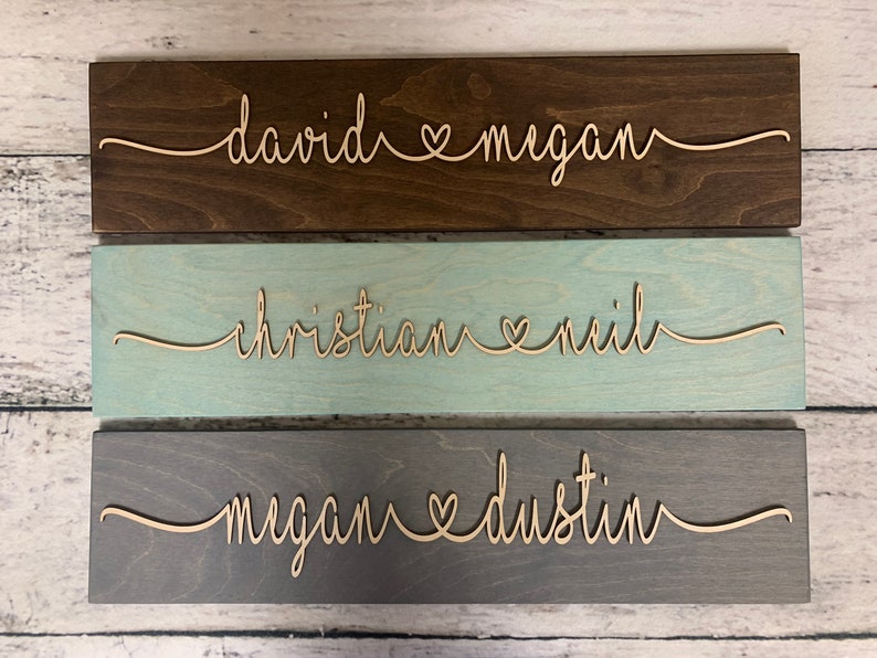 Puede incluir: Tres letreros de madera con nombres y un s&iacute;mbolo de coraz&oacute;n en escritura cursiva. El letrero superior es marr&oacute;n oscuro con los nombres "david" y "megan". El letrero del medio es azul claro con los nombres "christian" y "neil". El letrero inferior es gris con los nombres "megan" y "dustin".