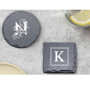 May include: Two slate coasters with personalized monograms. The first coaster features a decorative letter "N" with the name "NOLAN" below it. The second coaster features a letter "K" in a square frame.