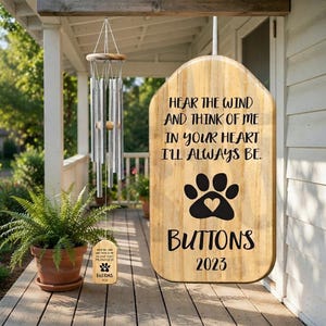 May include: A wooden wind chime featuring silver metal tubes and a rectangular wooden plaque with the text Hear the wind and think of me in your heart I'll always be, a black paw print with a heart center, the name Buttons, and the year 2023.