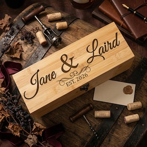 May include: A wooden wine box with the names "Jane & Laird" and "EST. 2026" engraved on the lid. The box is rectangular and has a metal clasp. Corkscrews and wine corks are scattered around the box on a wooden surface.