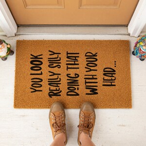 A textured door mat with a humorous message printed on it, placed on a tiled floor next to a plant.