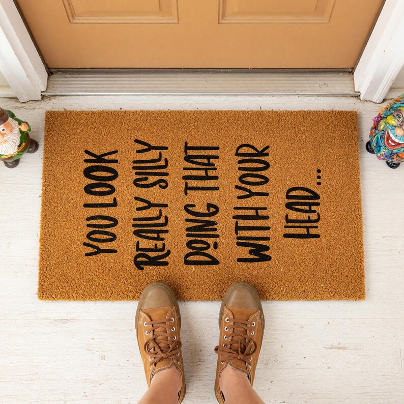 A textured door mat with a humorous message printed on it, placed on a tiled floor next to a plant.