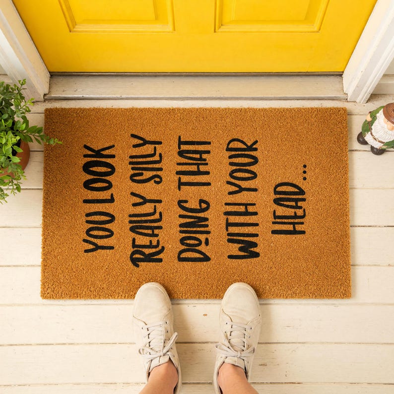A textured door mat with the phrase "You look really silly doing that with your head" printed on it, placed on a wooden surface with some greenery and red sandals.