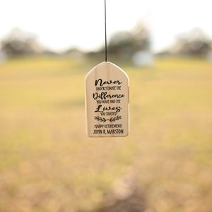 May include: A wooden sign with the text "Never underestimate the difference you made and the lives you touched. Happy Retirement! John R. Marston" hanging from a black string.