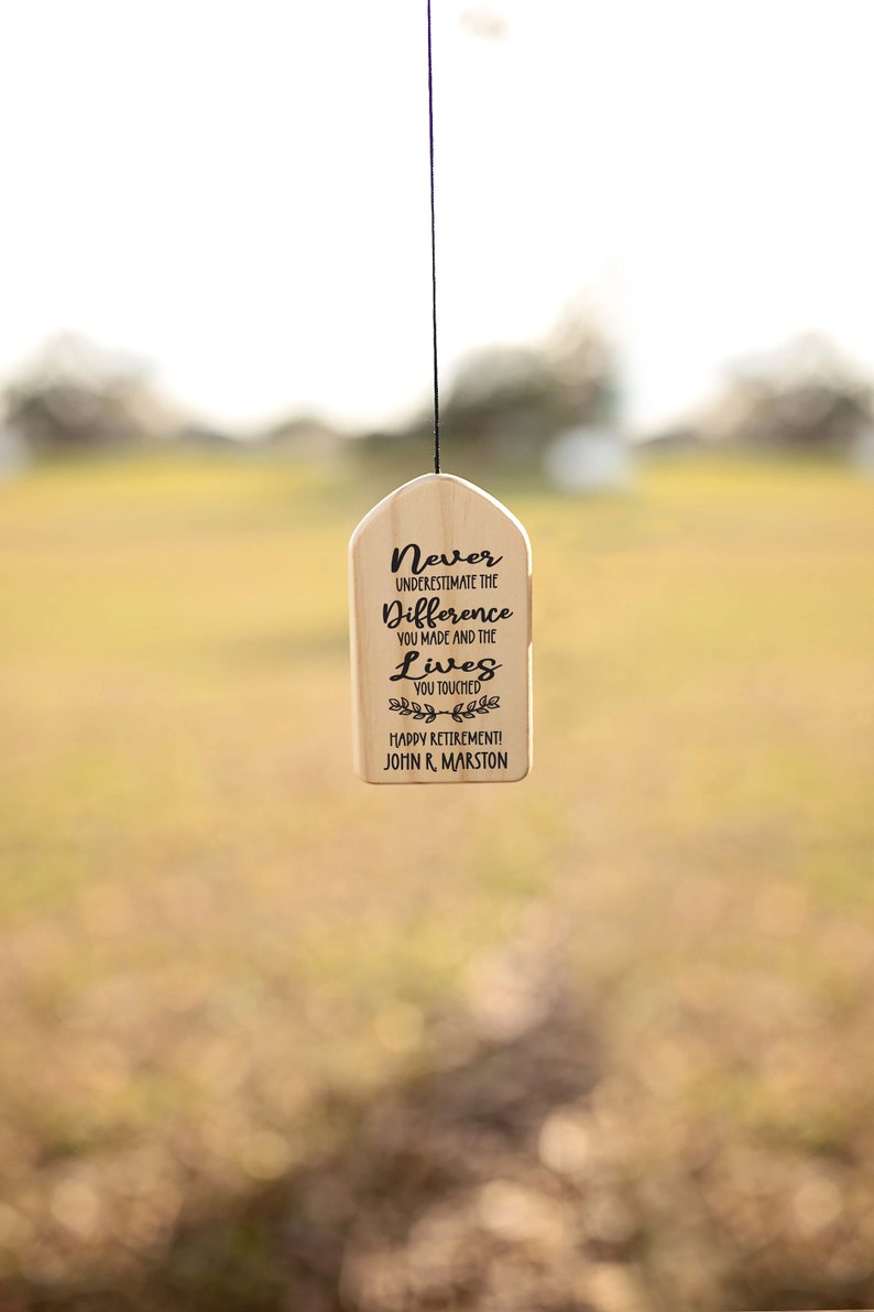 May include: A wooden sign with the text "Never underestimate the difference you made and the lives you touched. Happy Retirement! John R. Marston" hanging from a black string.