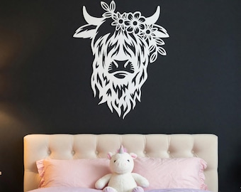 May include: A white wall-mounted cutout of a Highland cow head featuring long, shaggy fur and a floral crown between its horns. The design is rendered in a flat, solid white color with intricate cutouts defining the facial features and hair.