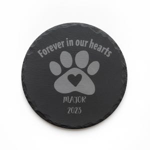 May include: A round black slate coaster with a rough, natural edge. The surface is engraved with a paw print containing a small heart, the text "Forever in our hearts", the name "MAJOR", and the year "2023" in a light gray color.